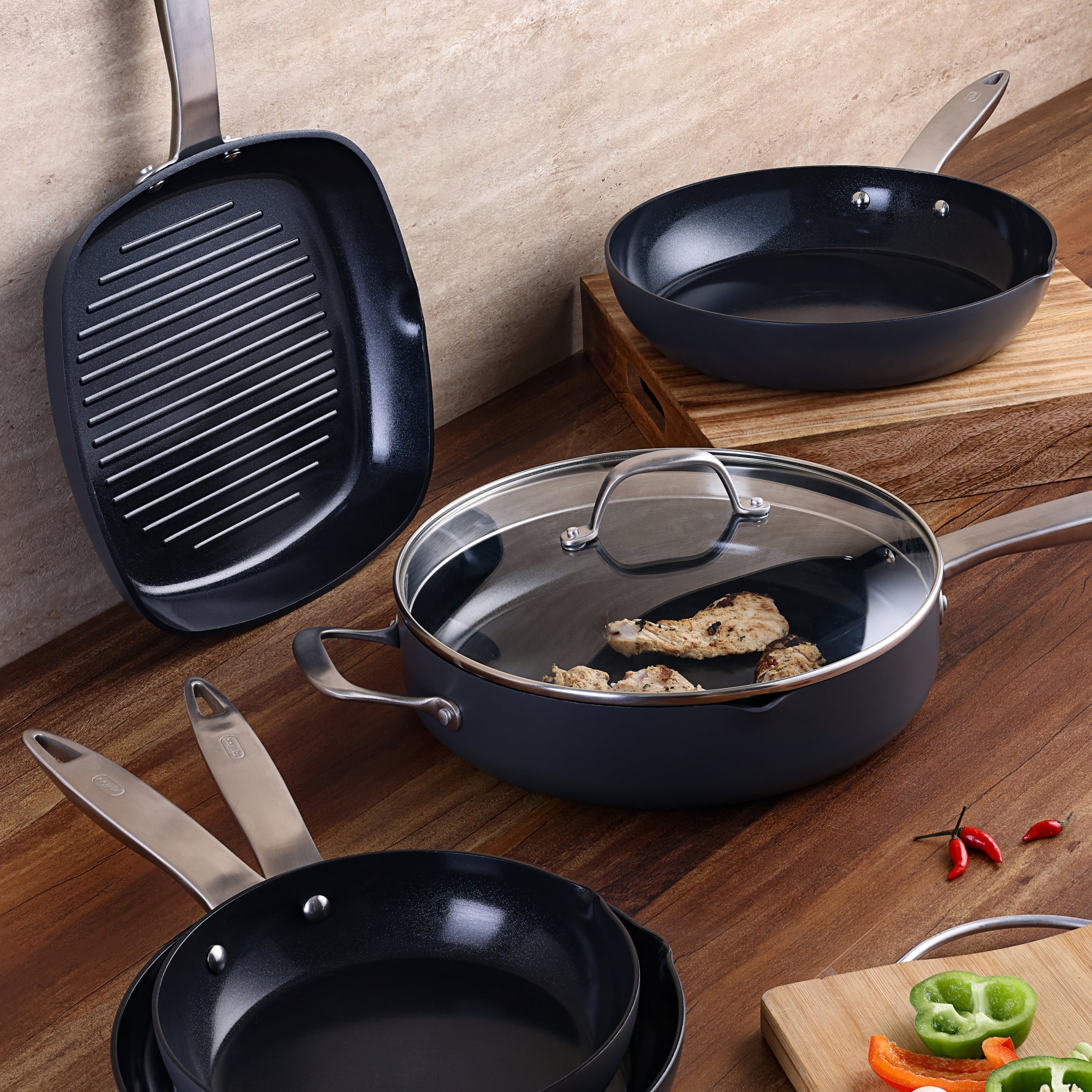 Four non-stick pans, including a grill pan and one with a glass lid containing cooked chicken pieces, are displayed on a wooden countertop near sliced vegetables.