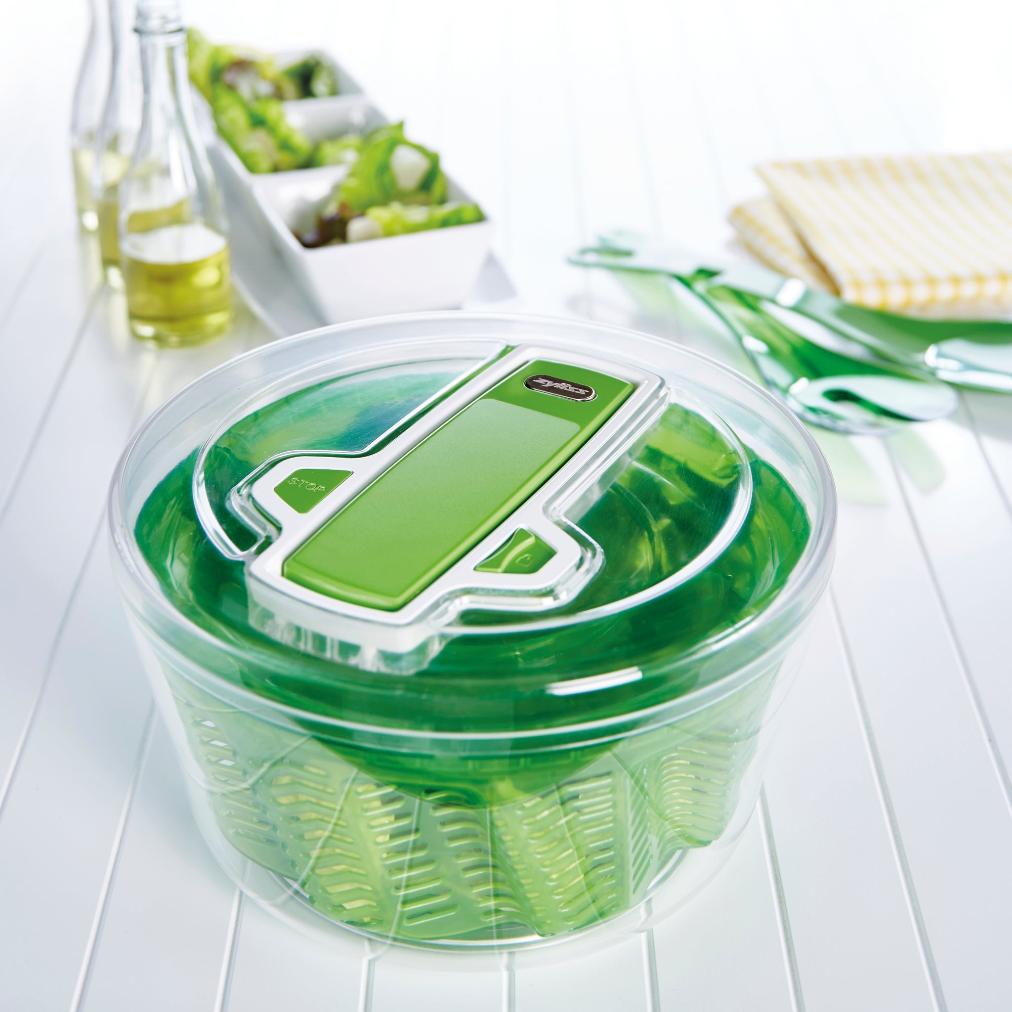 The Zyliss Swift Dry Salad Spinner Large, featuring a clear lid and non-slip base, rests on a white table. Behind it are green utensils, a yellow-striped cloth, olive oil, and a bowl of fresh vegetables.