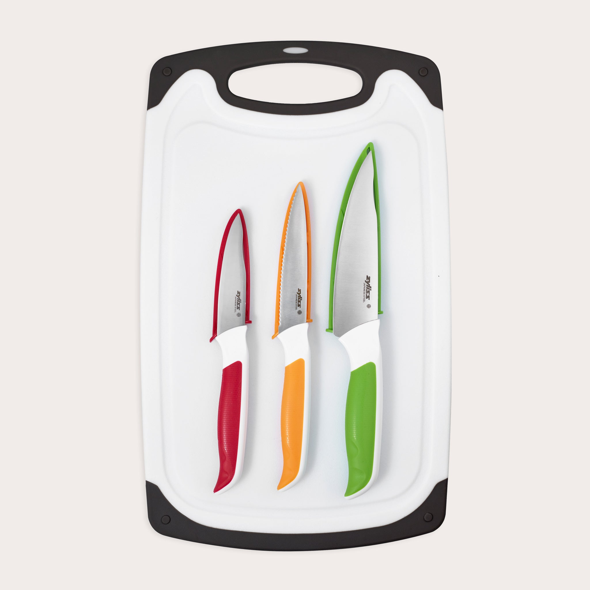 The Zyliss 4 Piece Comfort Board and Knife Set features a white board with black edges, holding three dishwasher-safe Japanese stainless steel knives with colored handles in red, orange, and green arranged from smallest to largest.