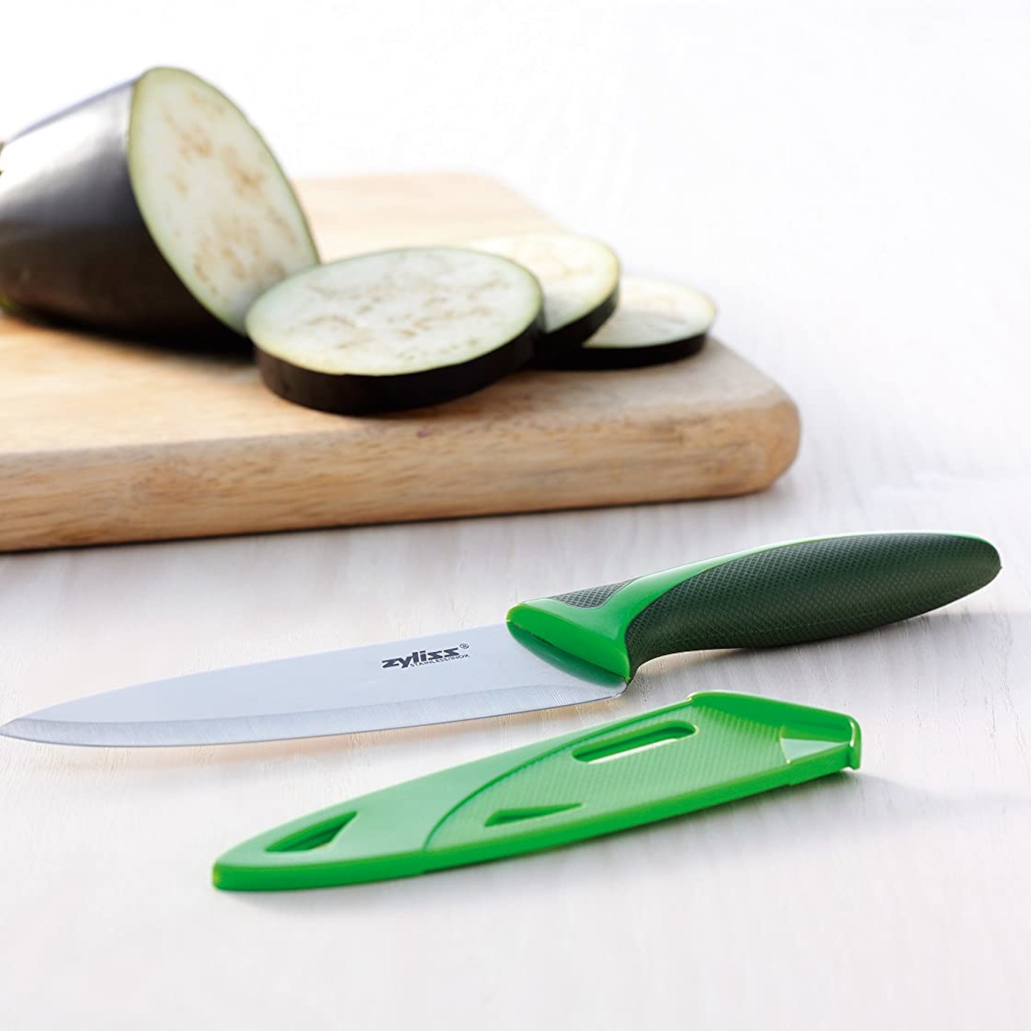 A green-handled kitchen knife with its matching blade cover lies on a white surface next to a cutting board with a partially sliced eggplant.