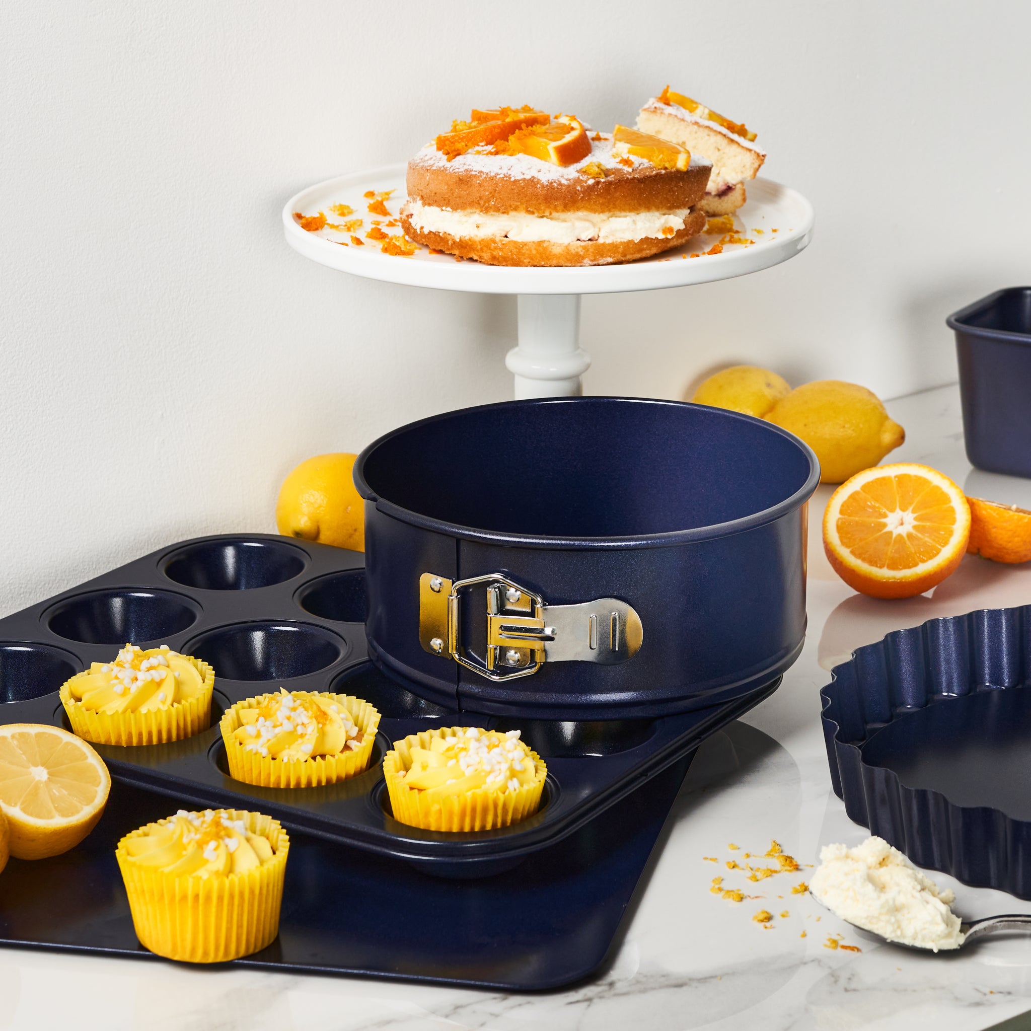A kitchen scene featuring the Zyliss Durable Non-Stick 12 Hole Muffin Pan, cupcakes, lemons, and oranges on a marble counter. In the background, a sliced cake sits on a white stand with scattered citrus fruits and zest.