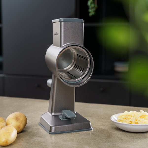 #drumgraterset  A Zyliss Gourmet Drum Grater sits on a kitchen counter, flanked by potatoes on the left and a plate of grated cheese on the right, with a softly blurred kitchen background.