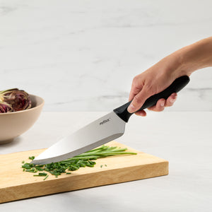A hand uses a Zyliss 6 Piece Knife Set to chop green herbs on a wooden board. Behind, a beige bowl with artichokes rests on a white surface, showcasing essential kitchen prep tools.