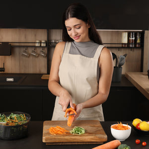 A woman in a beige apron smiles while using the Zyliss Julienne Y Peeler to slice a carrot over a wooden cutting board in a modern kitchen, with bowls of shredded vegetables and other ingredients on the counter.