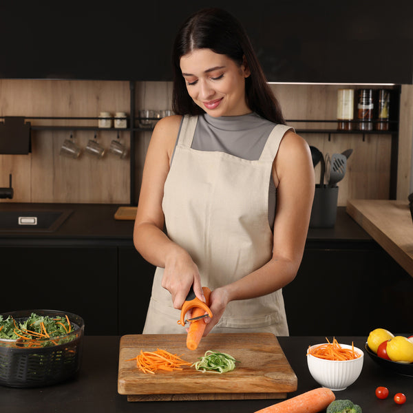 A woman in a beige apron smiles while using the Zyliss Julienne Y Peeler to slice a carrot over a wooden cutting board in a modern kitchen, with bowls of shredded vegetables and other ingredients on the counter.