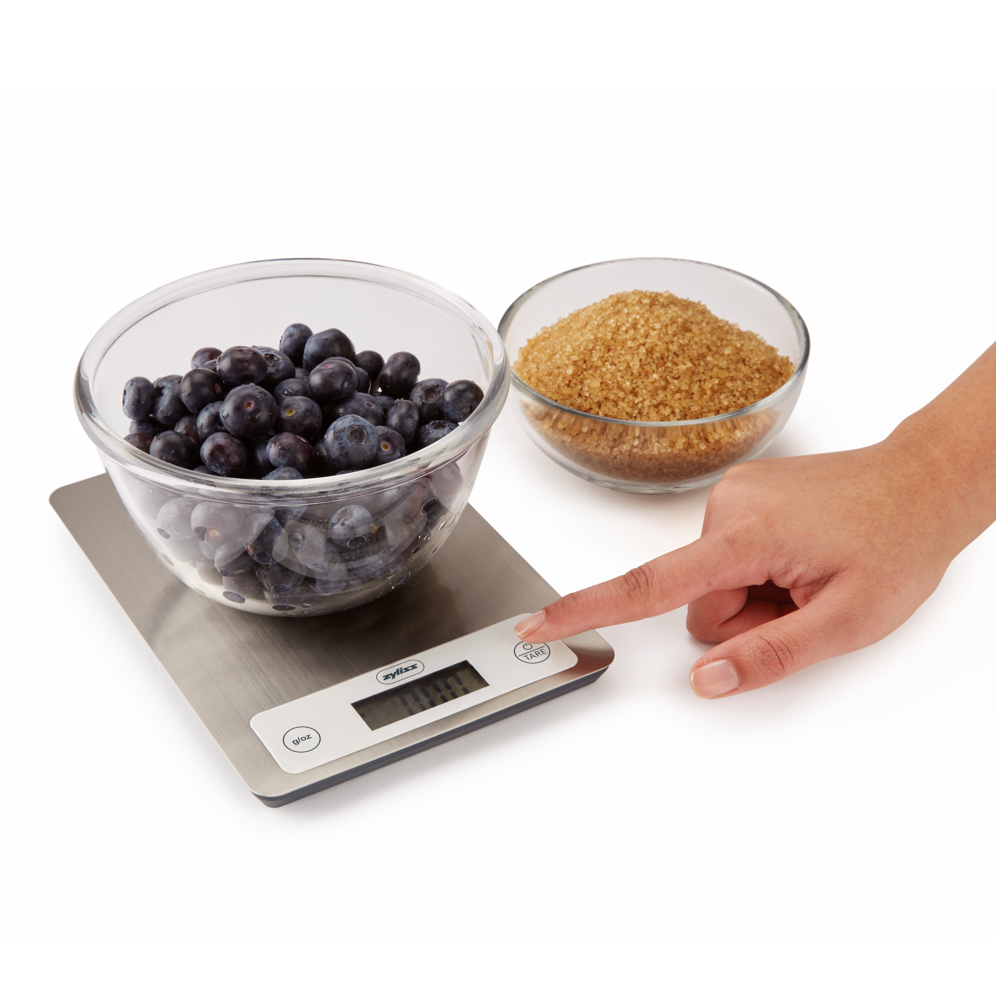 A hand presses the tare button on a digital kitchen scale with a bowl of blueberries on it. Next to the scale is a separate bowl filled with brown sugar.