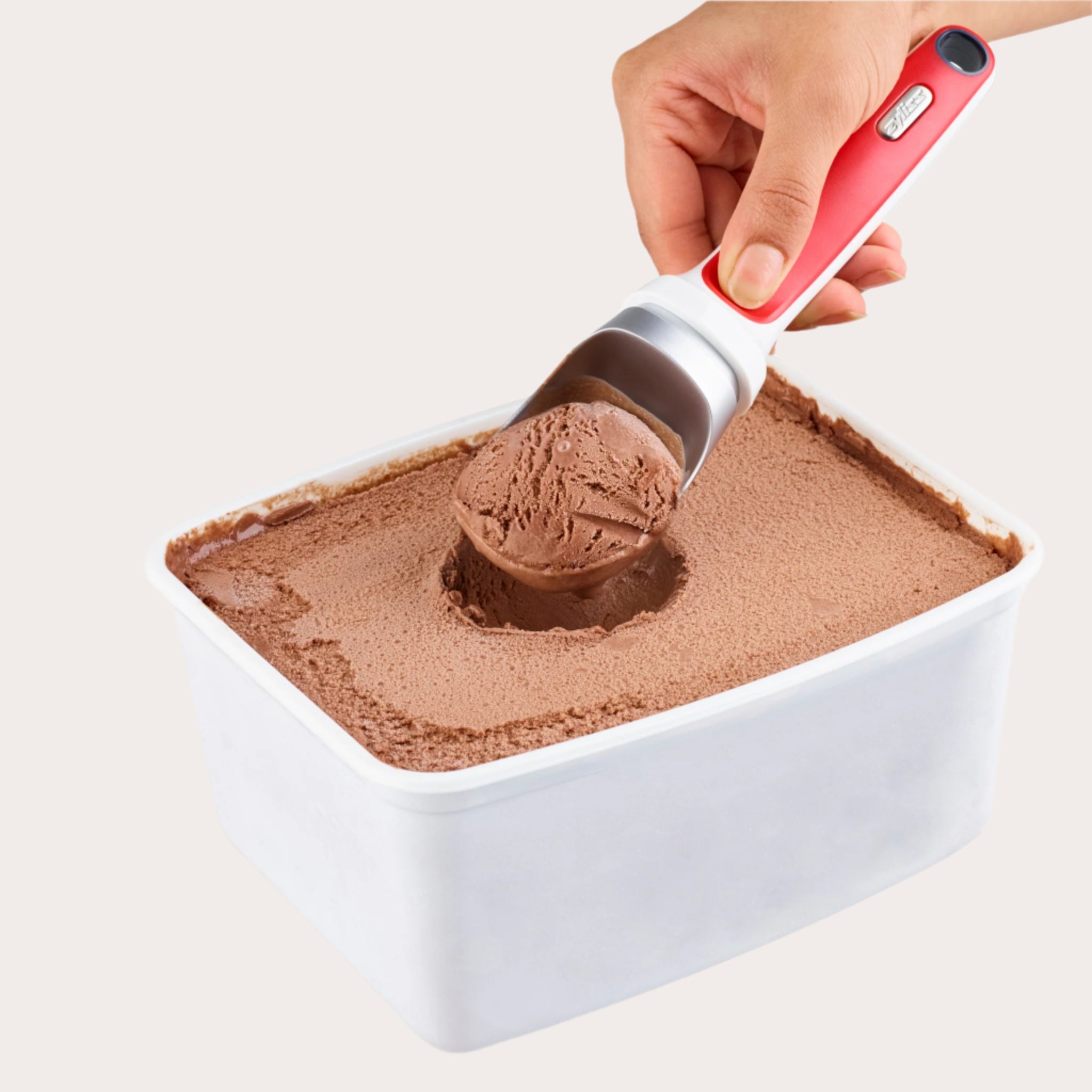 A hand uses the Zyliss Right Scoop Ice Cream Scoop, featuring a zinc alloy head and red ergonomic handle, to serve chocolate ice cream from a rectangular white container.