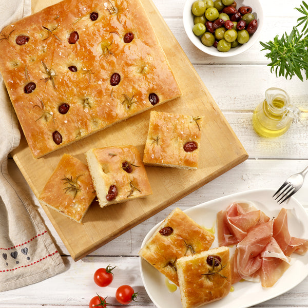 A Zyliss Durable Non-Stick Baking Tray holds olive and rosemary focaccia, served with prosciutto, mixed olives, cherry tomatoes, and olive oil—all beautifully arranged on a white rustic table.