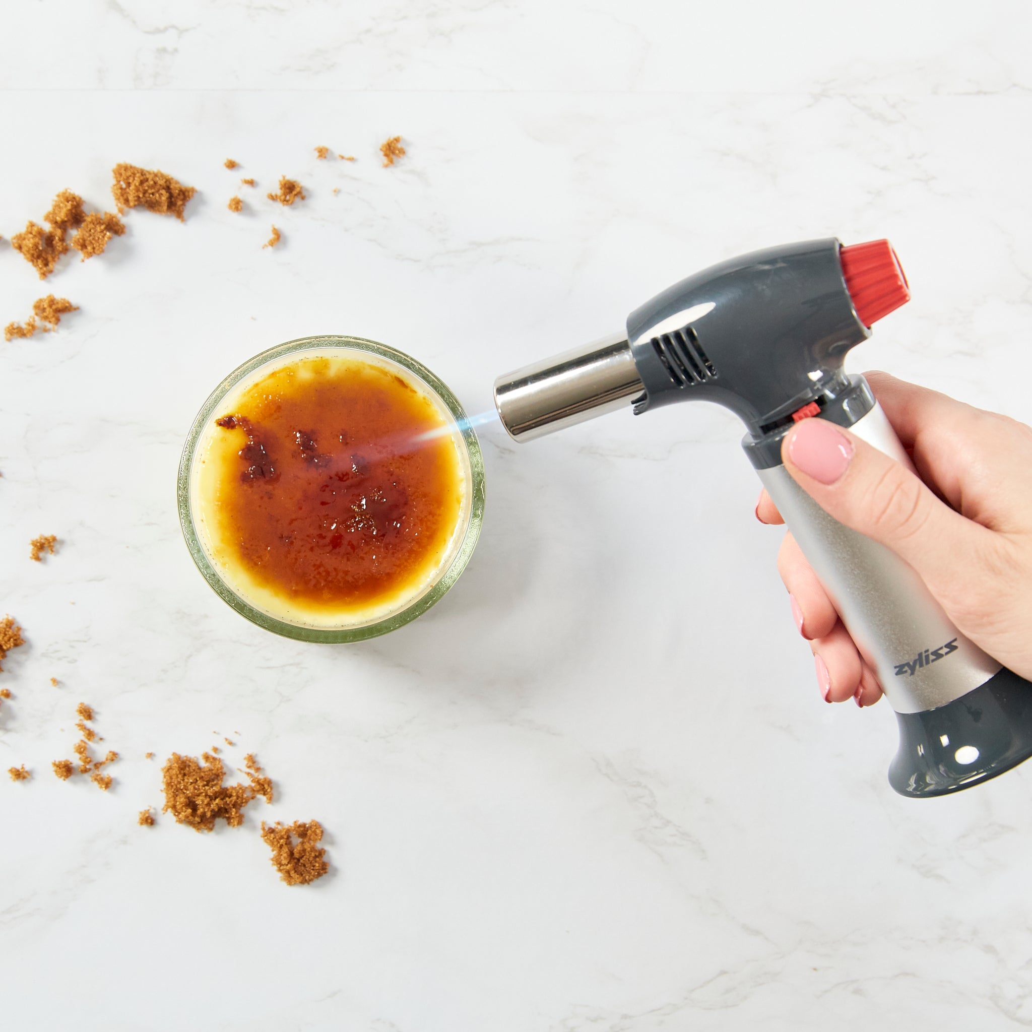A hand uses the Zyliss Kitchen Blowtorch, which features an ergonomic handle, to caramelize the sugar topping on a crème brûlée in a glass dish. Brown sugar crumbs are scattered on the marble surface nearby.