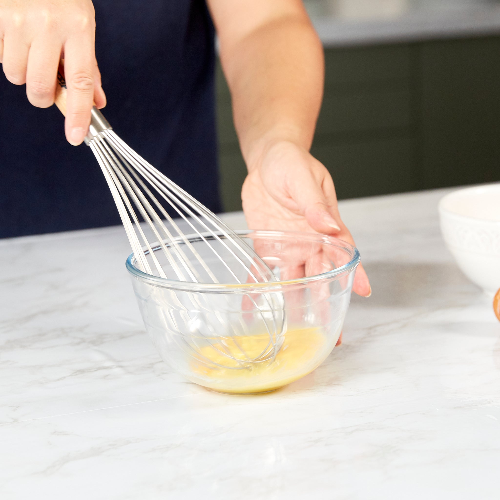 A person whisks eggs in a clear glass bowl on a white countertop, with part of another bowl visible in the background.