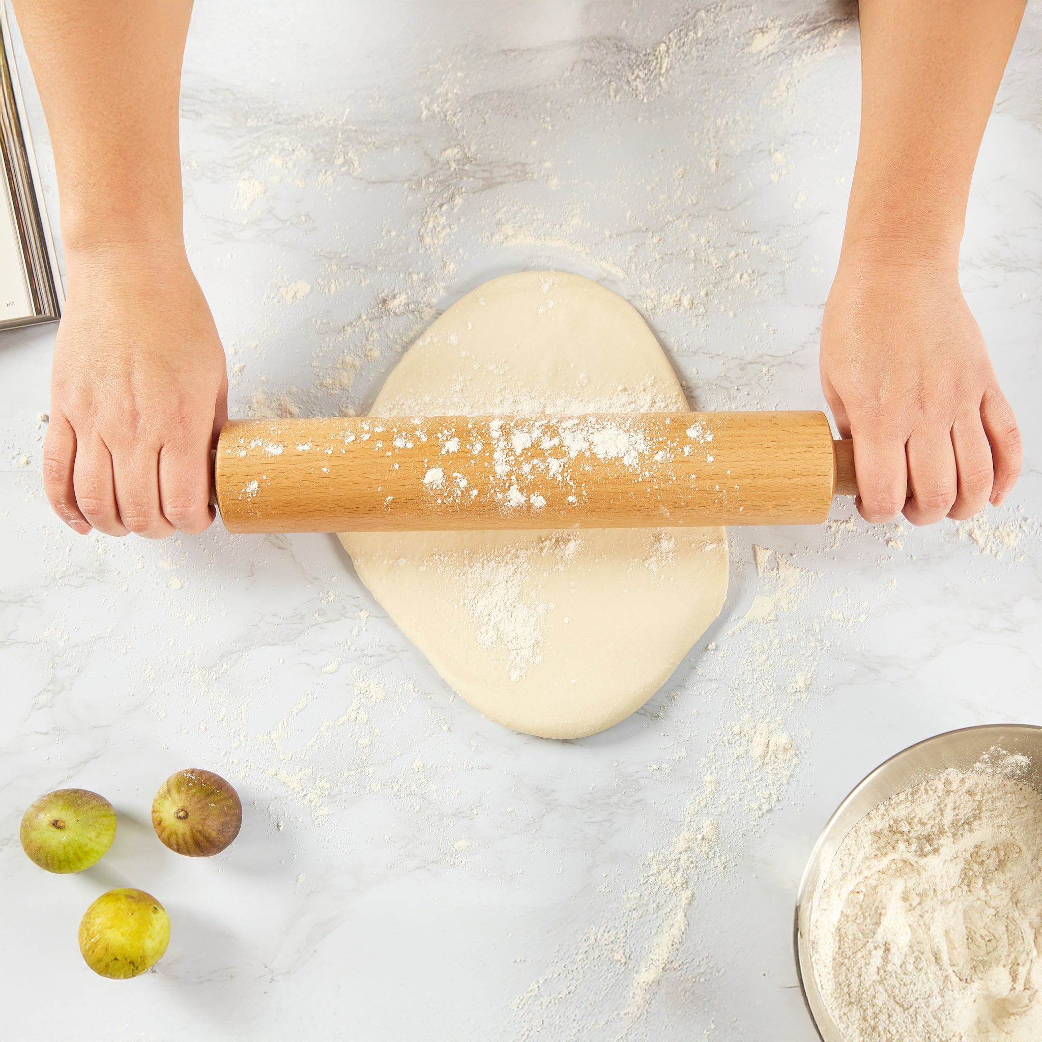 A person rolls out dough with a wooden rolling pin on a floured surface. Nearby are a bowl of flour and three figs.