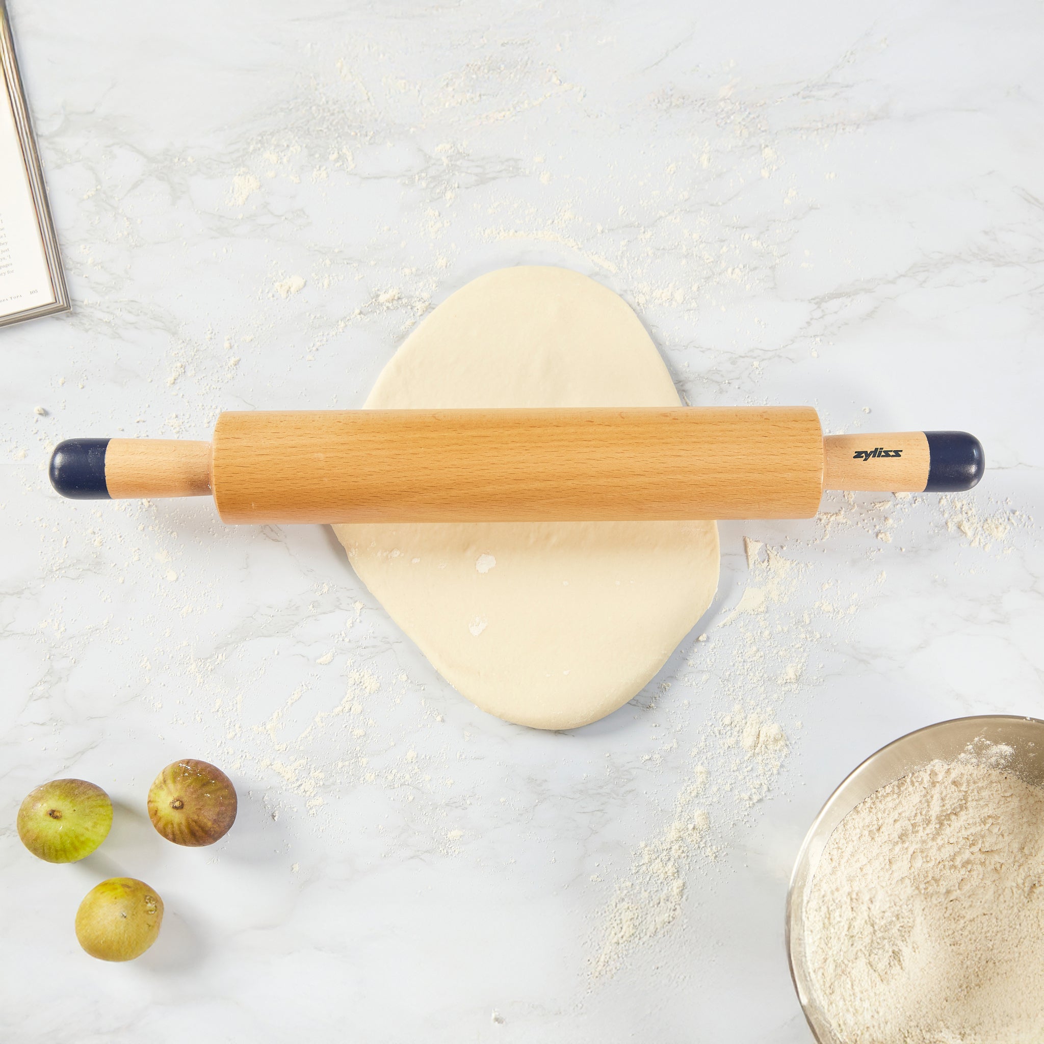 A Zyliss Beech Wood Rolling Pin with comfort-grip handles sits on dough on a floured surface, accompanied by three figs, a bowl of flour, and part of an open book—a must-have kitchen tool.