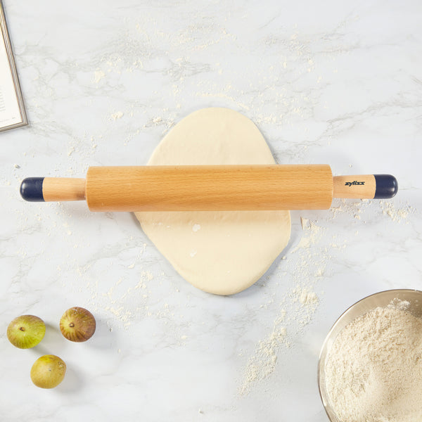A Zyliss Beech Wood Rolling Pin with comfort-grip handles sits on dough on a floured surface, accompanied by three figs, a bowl of flour, and part of an open book—a must-have kitchen tool.