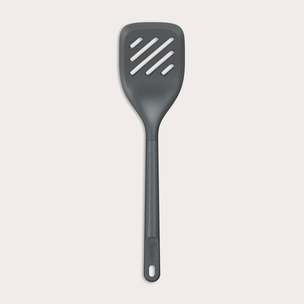 The Zyliss Slotted Turner, shown in black with three diagonal slots and an ergonomic handle, features a heat-resistant design for safe cooking or flipping food. Displayed against a plain white background.