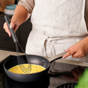 Wearing a light-colored apron, someone uses the Zyliss Flat Whisk Silicone to stir eggs in a nonstick frying pan on the stovetop.