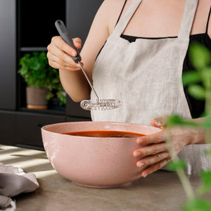 Wearing a light apron, a person uses the Zyliss Gravy Whisk to mix ingredients in a large pink bowl on the kitchen counter. Green plants and a towel appear in the background, enhancing the sauce preparation scene.