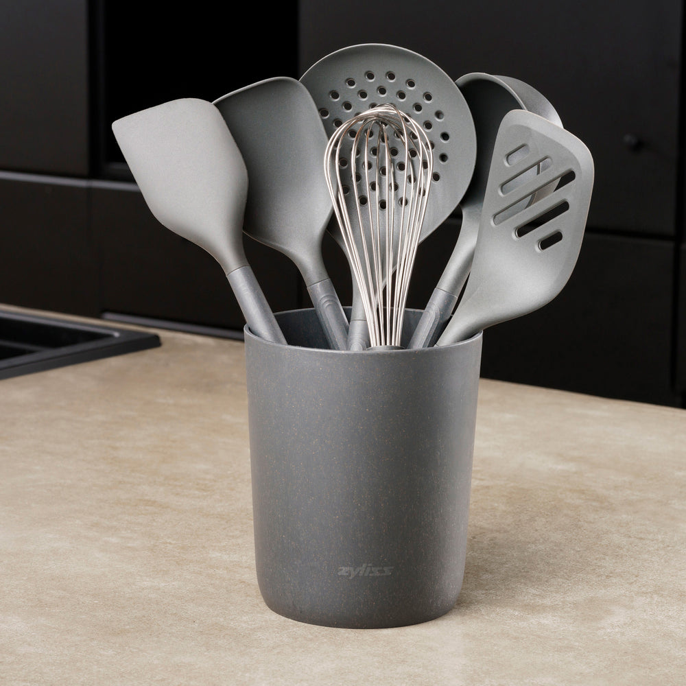 The Zyliss 7-Piece Utensil Set includes black kitchen tools—whisk, spatulas, spoon, and slotted spoon—organized in a matching container. Each features ergonomic handles and heat-resistant PBT for safe, comfortable use.