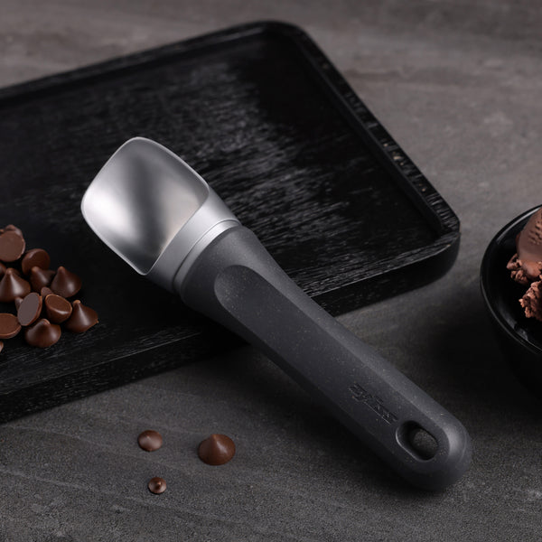 #belugagrey A Zyliss Ice Cream Scoop with an eco-friendly handle sits on a textured black tray beside chocolate chips and a bowl of chocolate ice cream, arranged on a gray surface.
