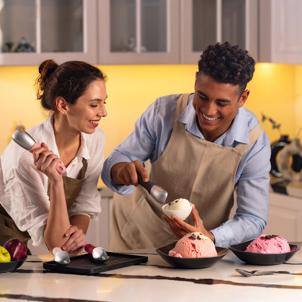 #naturalbeige A man and woman in aprons smile as they use the Zyliss Ice Cream Scoop, featuring a sustainable wheat straw, eco-friendly handle, to serve various flavors into bowls on the counter.