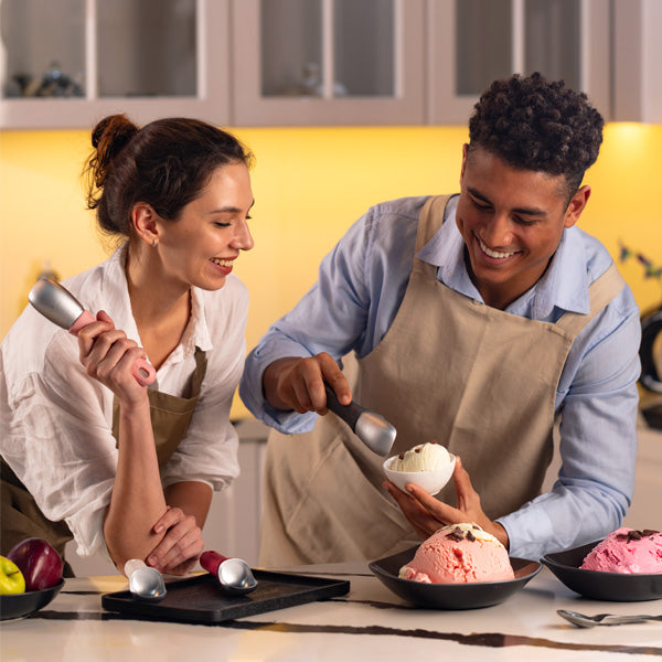 A smiling man and woman wearing aprons are scooping ice cream in a kitchen. The man serves vanilla ice cream into a bowl while the woman looks on, holding an ice cream scoop. Bowls of ice cream are on the counter.