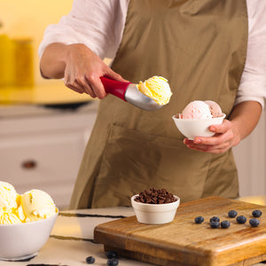 #berry Wearing a beige apron, a person uses the Zyliss Ice Cream Scoop to serve vanilla ice cream into a small bowl. Other bowls of ice cream, chocolate chips, a cutting board, and scattered blueberries are on the counter.