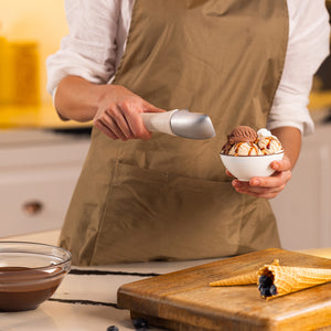 #naturalbeige Wearing a brown apron, a person uses a Zyliss Ice Cream Scoop with an eco-friendly handle to serve ice cream into a syrup-topped bowl beside empty sustainable wheat straw waffle cones and melted chocolate.