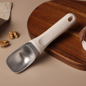 #naturalbeige A Zyliss Ice Cream Scoop with an eco-friendly handle sits on a round wooden board, surrounded by walnuts, with a bowl of chocolate ice cream partially visible.
