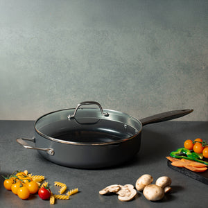 A Zyliss Ultimate Pro Ceramic Saute Pan With Lid rests on a gray counter, with uncooked pasta, cherry tomatoes, mushrooms, sliced carrots, and green bell peppers around it.