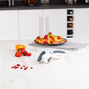 A plate of assorted fresh fruit sits on a folded napkin on a white kitchen counter, with the Zyliss Lock & Lift Can Opener, strawberries, an orange slice, and scattered pomegranate seeds in the foreground.