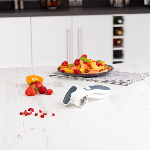 A plate of assorted fresh fruit sits on a folded napkin on a white kitchen counter, with the Zyliss Lock & Lift Can Opener, strawberries, an orange slice, and scattered pomegranate seeds in the foreground.