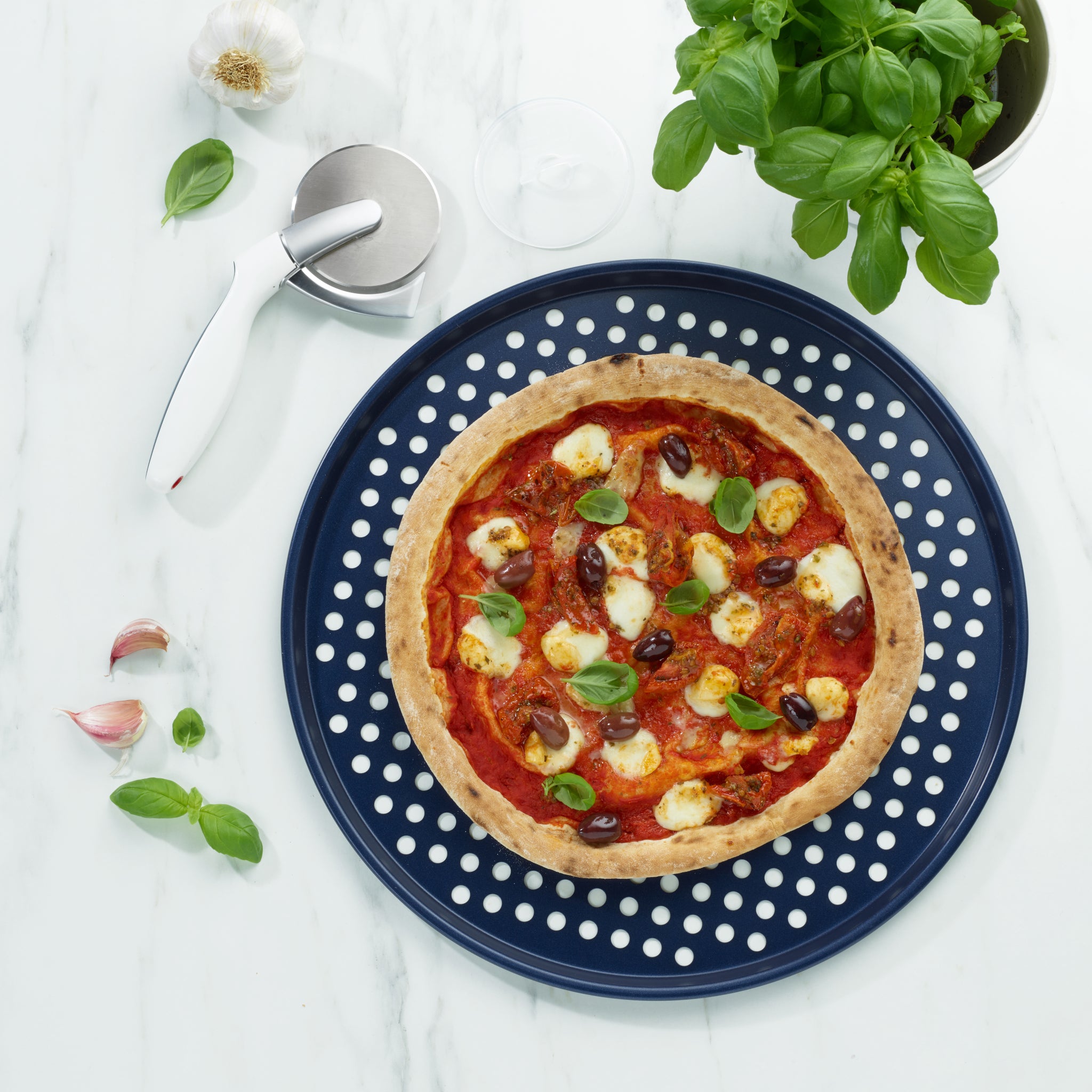 A pizza with mozzarella, olives, and basil rests on a blue polka dot plate. A Zyliss Durable Non-Stick Pizza Tray sits nearby on white marble, alongside a pizza cutter, fresh basil, garlic cloves, and a glass.