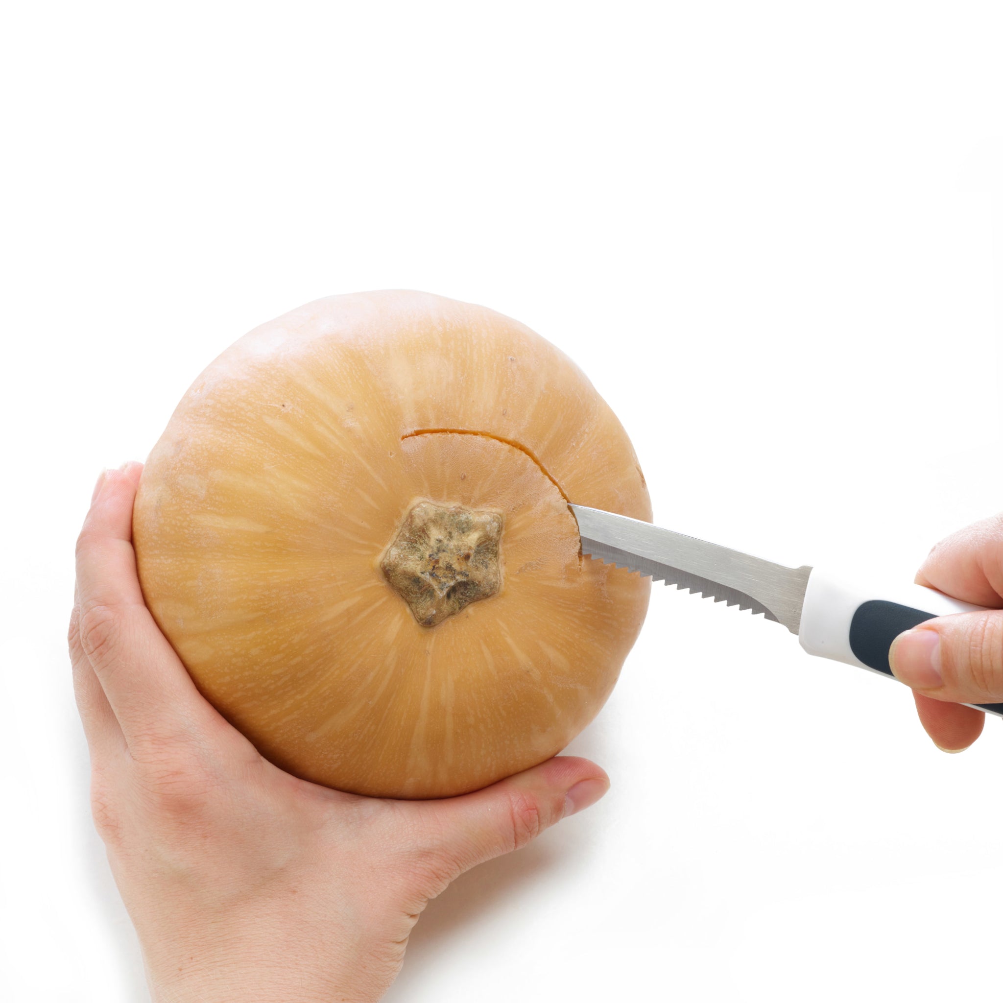 A person holds a round pumpkin while cutting into the top near the stem with a serrated knife against a white background.