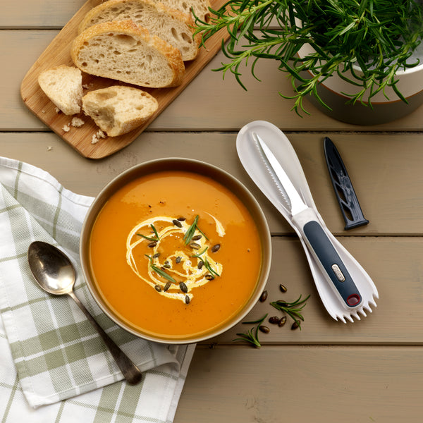 A bowl of creamy orange soup sits on a wooden table, garnished with cream, seeds, and herbs. Its made using the Zyliss 3 In 1 Pumpkin & Squash Scoop & Knife. Nearby are bread slices, a serrated knife, cutlery, napkin, and potted rosemary.