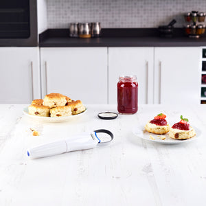 On a white kitchen counter sit scones and red jam. Nearby rests the Zyliss StrongBoy 2 Jar Opener, featuring an adjustable grip and non-slip design, adding function to this modern kitchen scene.