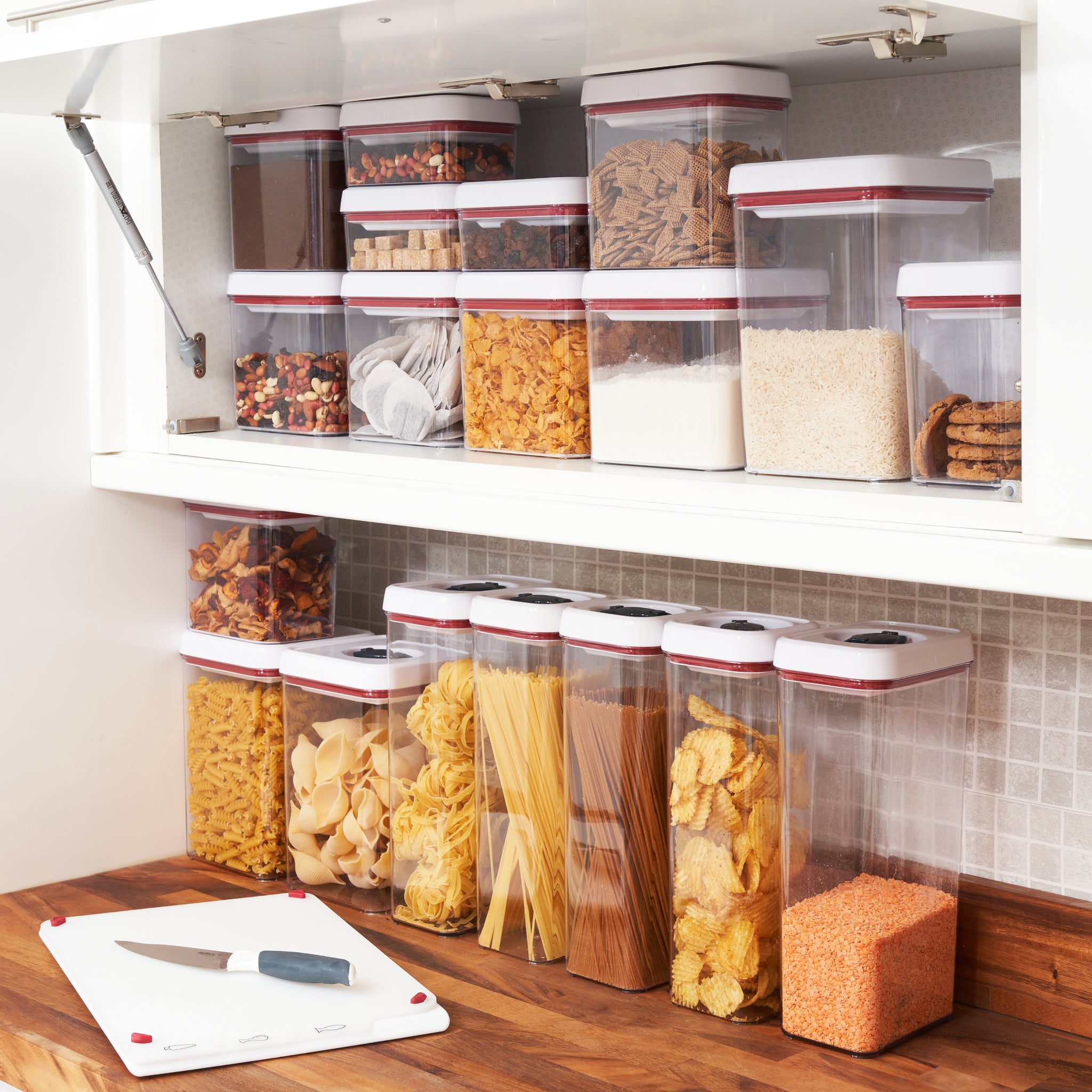 Clear containers with white lids neatly hold various dry foods like pasta, rice, cereals, and nuts on white shelves above a kitchen counter with a cutting board and knife.