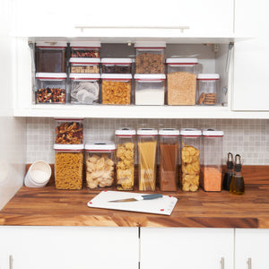 A kitchen countertop features the Zyliss Twist & Seal 5 Piece Container Set, efficiently storing dry foods like pasta, cereal, and nuts. Clear airtight containers with twist-lock lids, a cutting board, knife, olive oil bottles, and white canisters complete the scene.