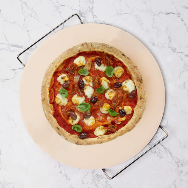 A homemade pizza with tomato sauce, mozzarella, black olives, and basil sits on the Zyliss Ceramic Baking Stone atop marble, featuring a golden crust.