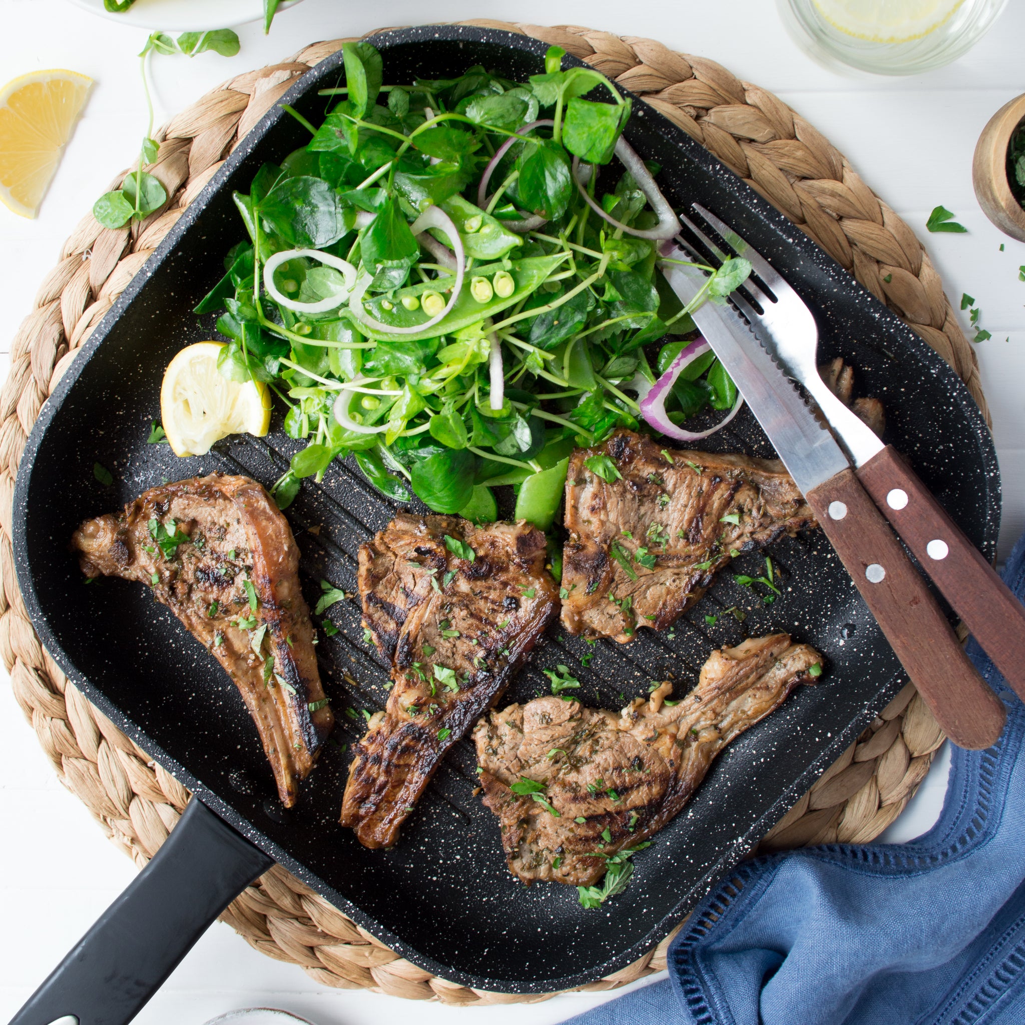 Grilled lamb chops with herbs on the Zyliss Ultimate Non-Stick Grill Pan, paired with a spinach, onion, and pea salad. A fork and knife rest on the pan, which is placed on a woven placemat.