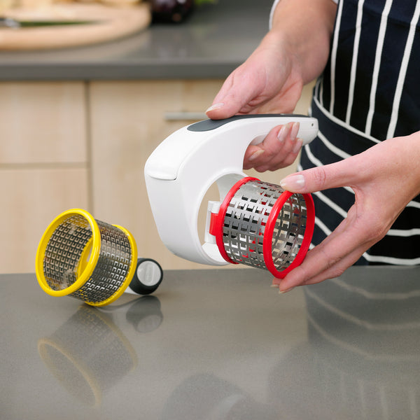 A person in an apron holds the All Cheese Grater by Zyliss, featuring sharp stainless steel blades and a white body with a red interchangeable drum, while a yellow drum attachment sits on the kitchen counter.