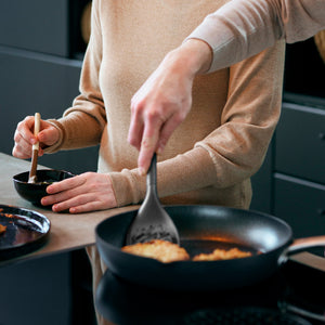 Two people cook together; one stirs a small bowl, while the other uses the Zyliss Slotted Turner with an ergonomic handle to cook food in a frying pan on the stove. Their faces arent visible.