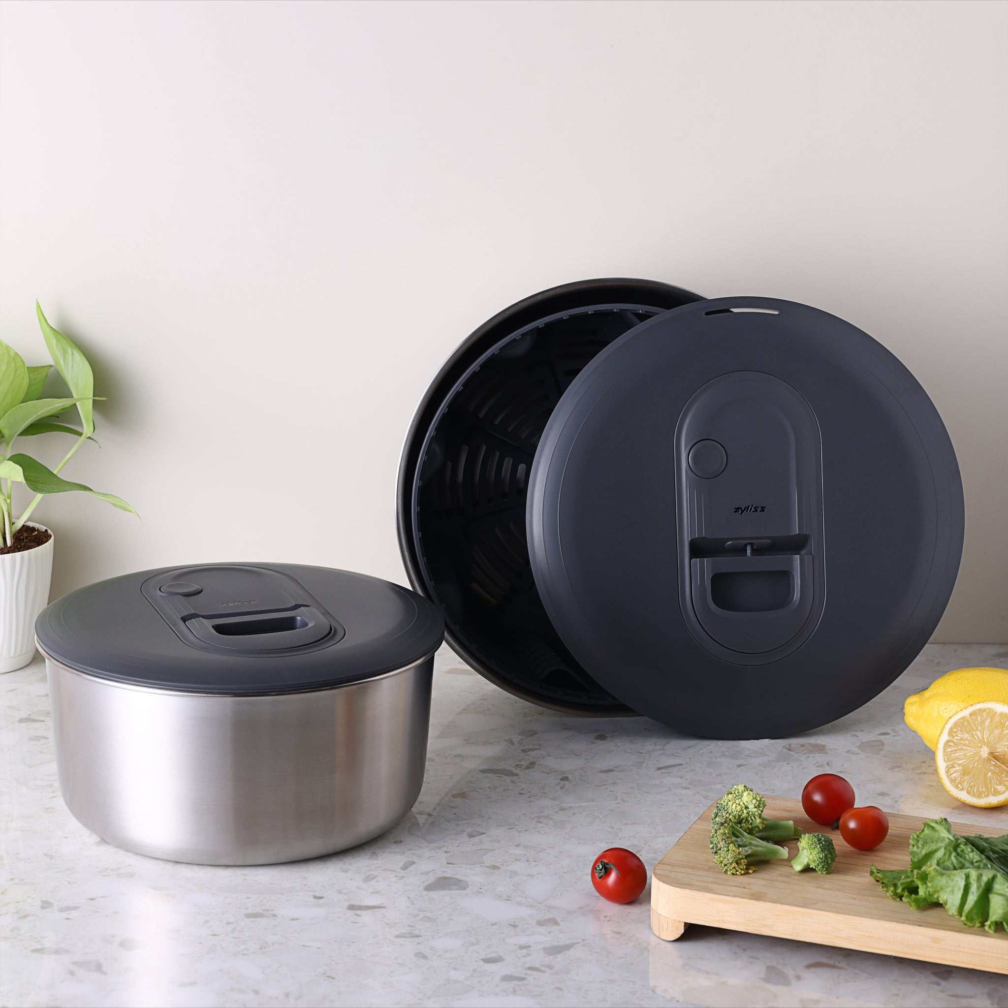 Two black-lidded containers are on a countertop, one open and one closed, with fresh vegetables, a lemon, and a potted plant nearby, set against a light, neutral background.