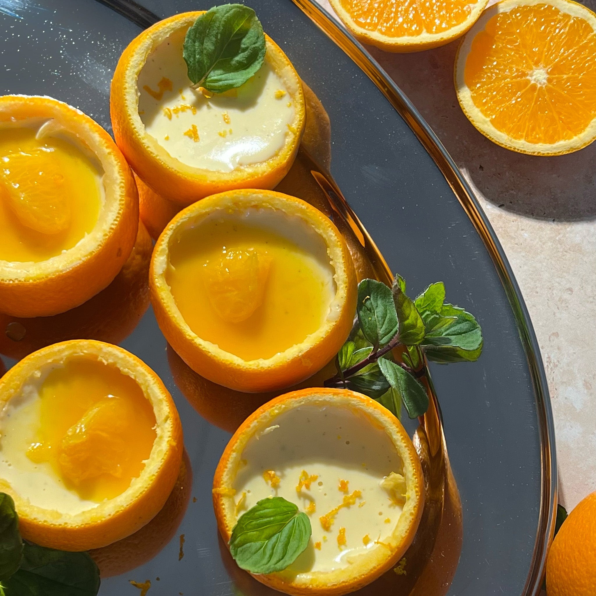 Four orange halves filled with creamy custard, garnished with orange segments and fresh mint leaves, are arranged on a reflective tray with two additional orange halves nearby.