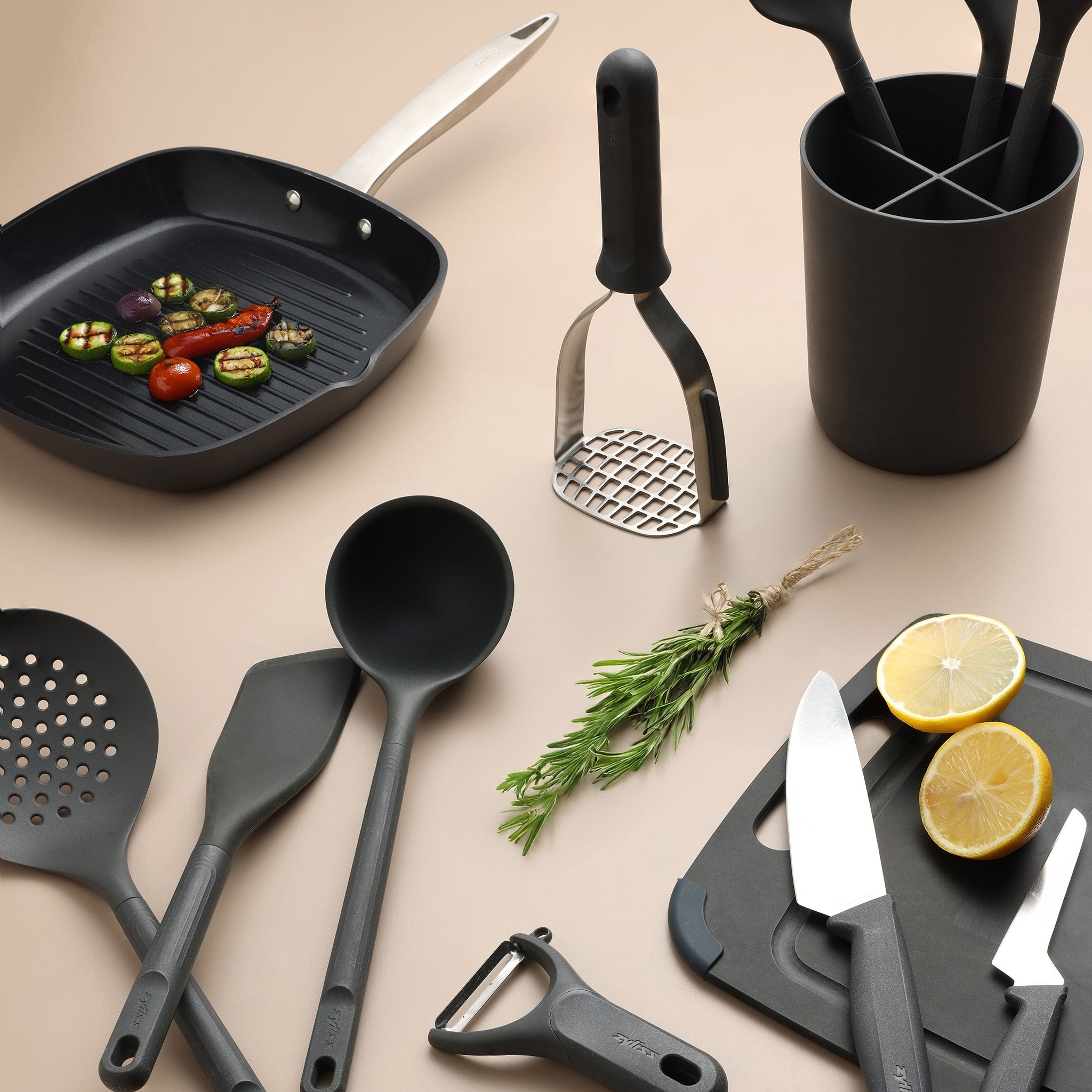 A selection of black kitchen utensils, knives, and a potato masher are arranged on a beige surface with a grill pan, sliced lemon, herbs, and vegetables.