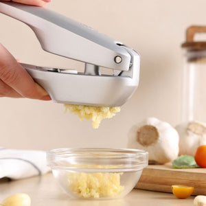 A hand uses the Zyliss Susi 4 Garlic Press with ergonomic handles to mince garlic into a glass bowl on a kitchen counter, with garlic bulbs and a yellow tomato in the background for easy cleanup.
