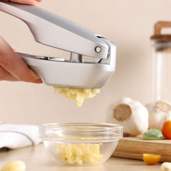 A hand uses the Zyliss Susi 4 Garlic Press with ergonomic handles to mince garlic into a glass bowl on a kitchen counter, with garlic bulbs and a yellow tomato in the background for easy cleanup.