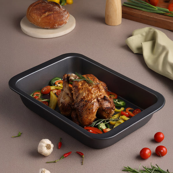 A roasted chicken rests in the Zyliss Non-Stick Oven Tray with sliced bell peppers and herbs, surrounded by fresh bread, cherry tomatoes, chili peppers, garlic, and a green napkin on a beige countertop.