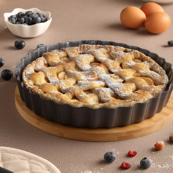 A freshly baked pie rests in the Zyliss Non-Stick Tart Pan with removable base on a wooden board, surrounded by blueberries, eggs, and scattered berries on a beige surface. A small bowl of blueberries is in the background.