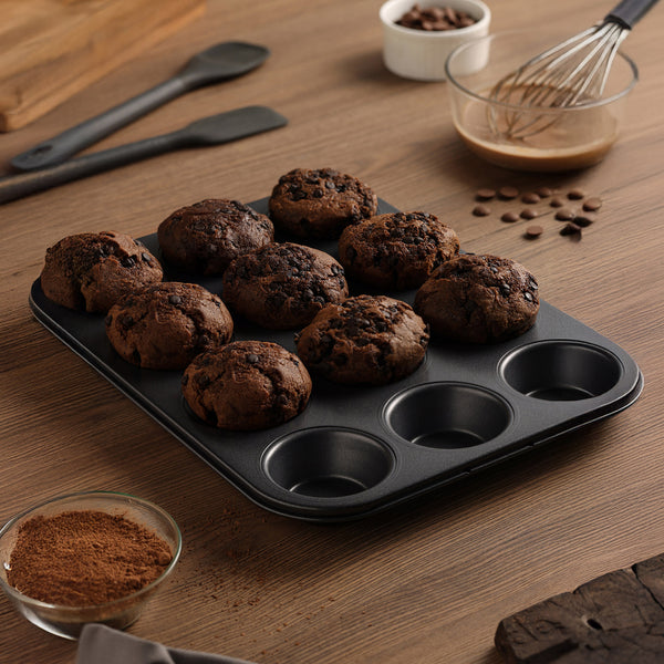 A Zyliss Non-Stick 12 Hole Muffin Pan with nine chocolate muffins rests on a wooden table, surrounded by baking tools, cocoa powder in a bowl, a whisk, chocolate chips, and a spatula.