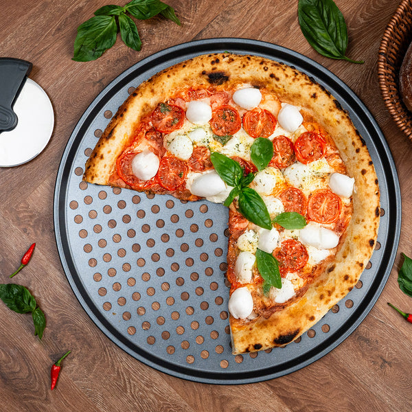 A Margherita pizza with tomato, mozzarella, and basil sits on a Zyliss Non-Stick Pizza Tray made of carbon steel, ensuring crispy bases. Basil, chili peppers, and a pizza cutter lie nearby on the wooden surface.