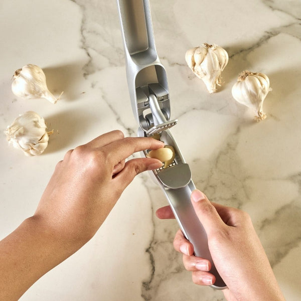 A person uses the Zyliss Susi 4 Garlic Press with ergonomic handles to press a clove on a marble countertop, surrounded by whole garlic bulbs, ensuring easy cleanup after use.